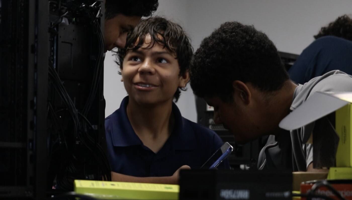 Student grinning mid-build, surrounded by classmates working inside a PC case